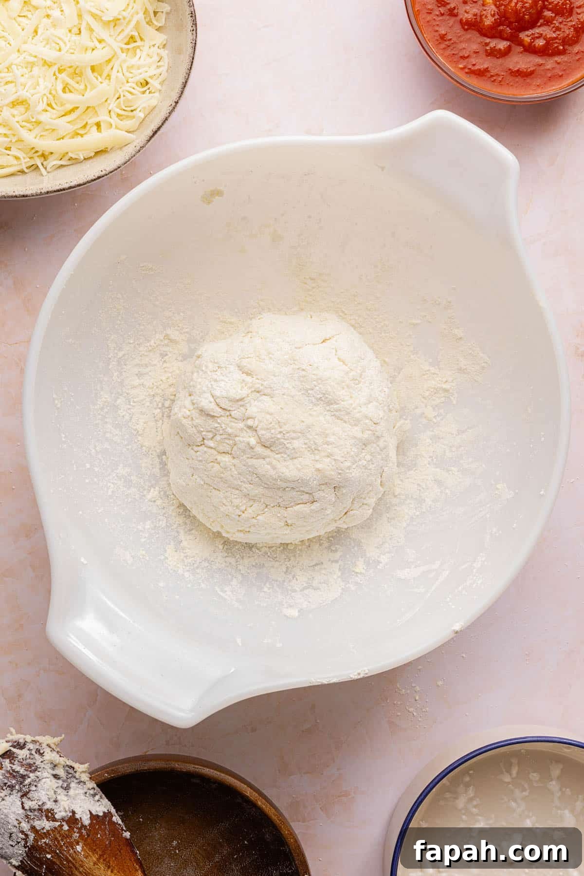 The pizza dough formed into a ball in a white bowl on a countertop.