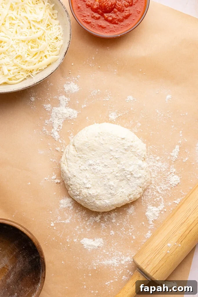 The pizza dough ball on a piece of brown parchment paper sprinkled with flour. A rolling pin sits off to the right side.