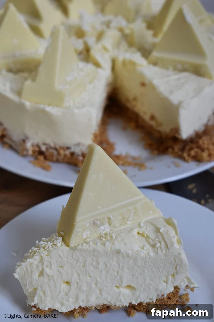 Vertical view of a slice of White Chocolate Toblerone Cheesecake on a white plate with the full cheesecake in the background on a brown board.
