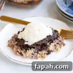 German Chocolate Dump Cake Recipe — Quick Decadent Dessert 7 A serving of chocolate and coconut dump cake topped with a scoop of vanilla ice cream on a white plate with a gold fork and blue plates in the background.