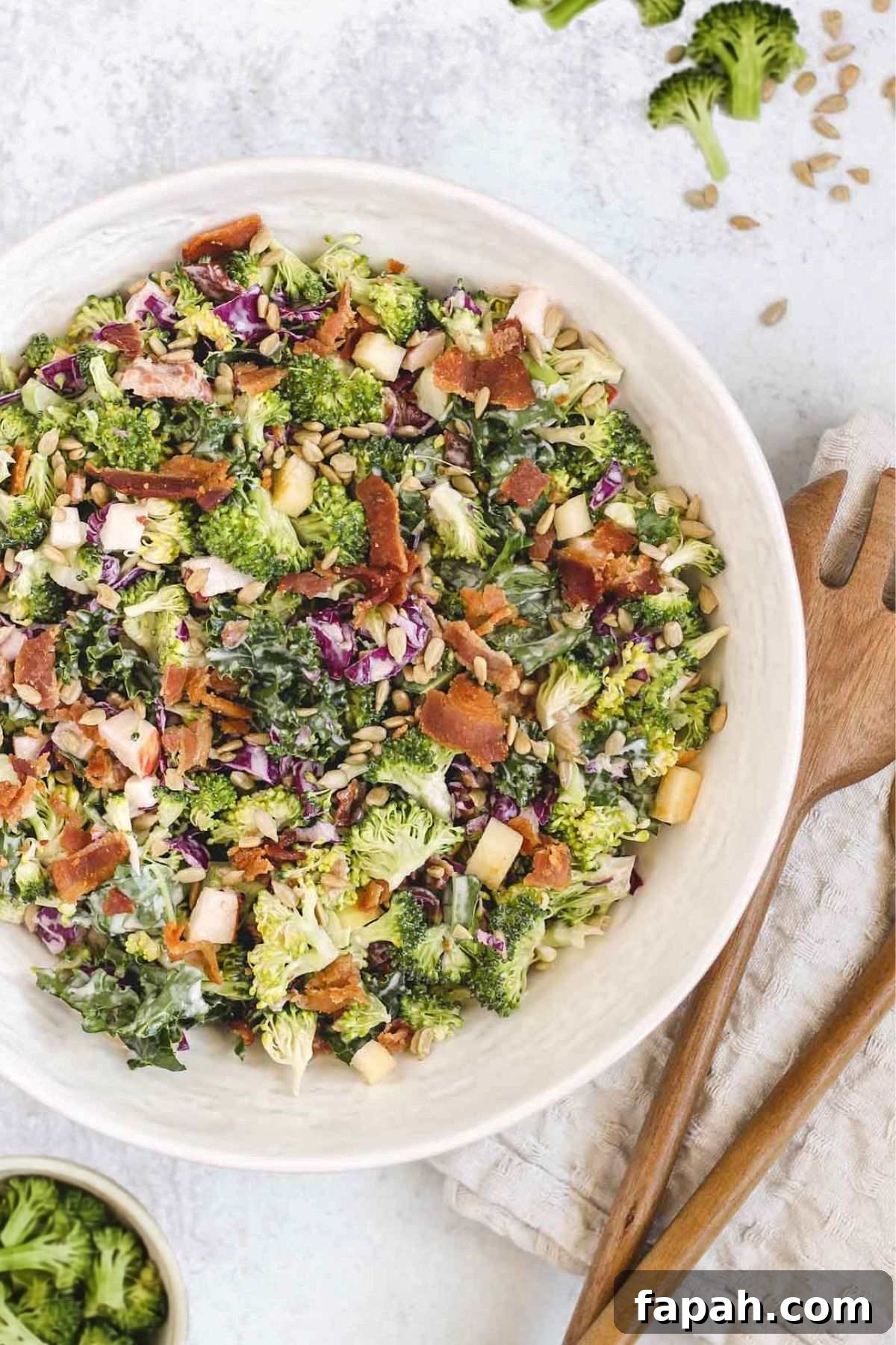Broccoli salad in a large serving bowl with wooden salad utensils laying next to the bowl and broccoli sprinkled around the bowl.