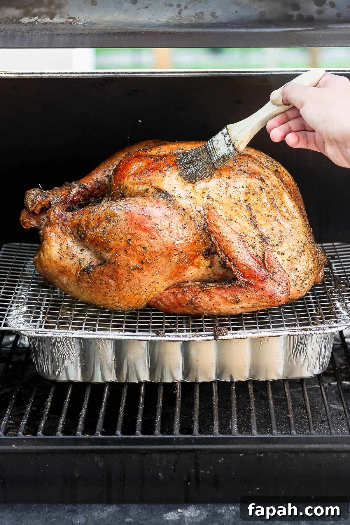 A whole turkey being cooked on a smoker with someone basting it with a brush. 