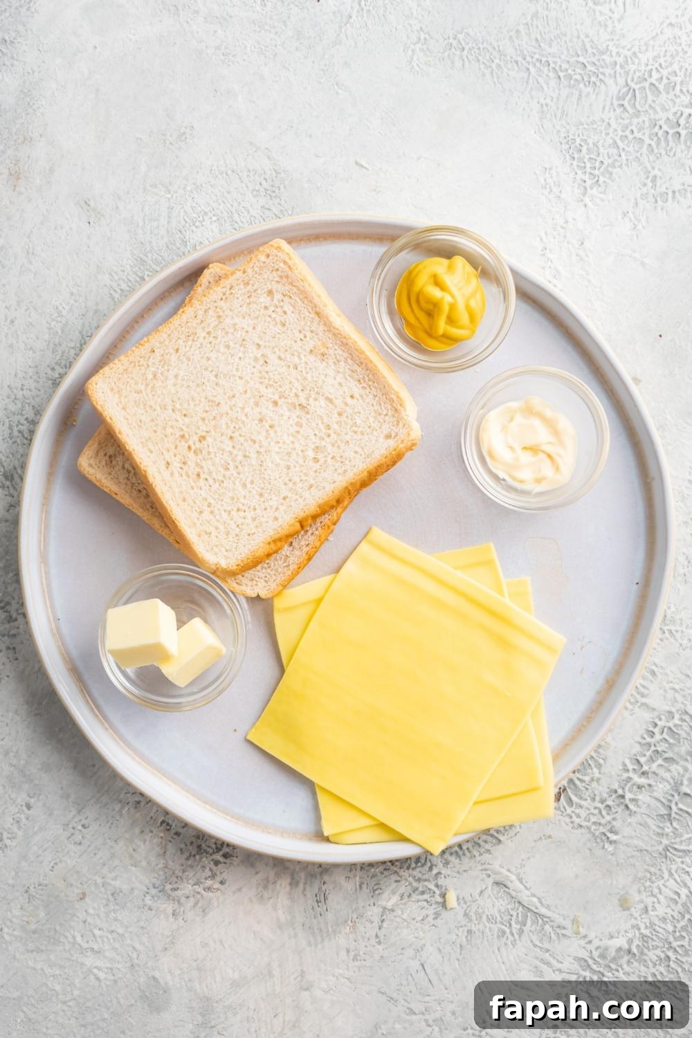An overhead view of two slices of white bread, three slices of cheese, and three small bowls containing mustard, mayo, and butter.