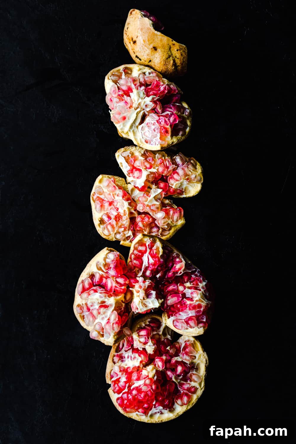 A white pomegranate with pink arils, cut into pieces and arranged vertically; overhead shot on a black background.
