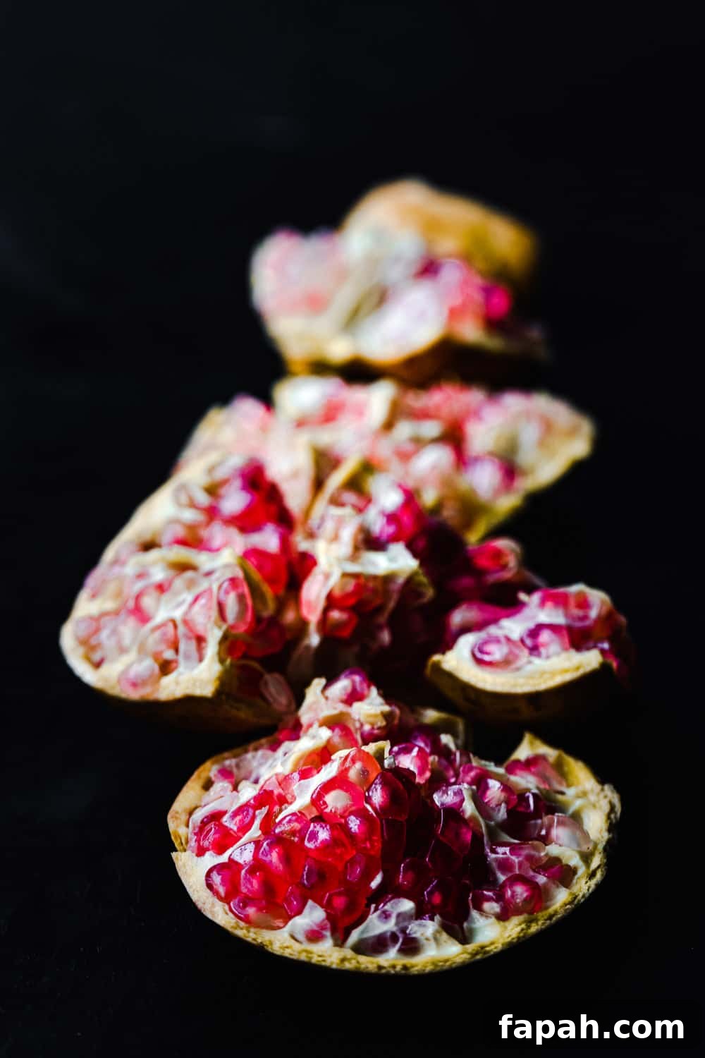 Side angle shot of a white pomegranate cut into pieces with the pink arils exposed, on a black background.