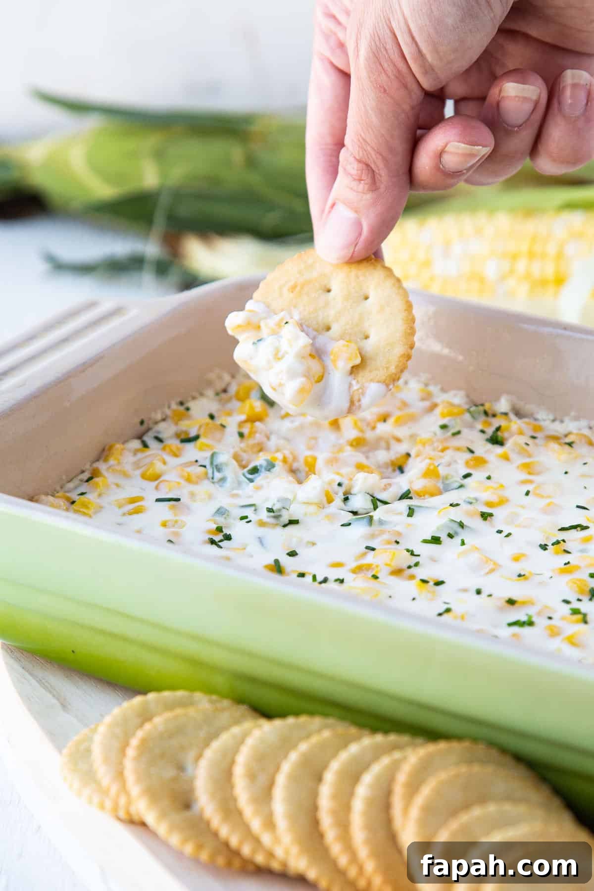 hand dipping a cracker into cream cheese corn dip in a green baking dish.