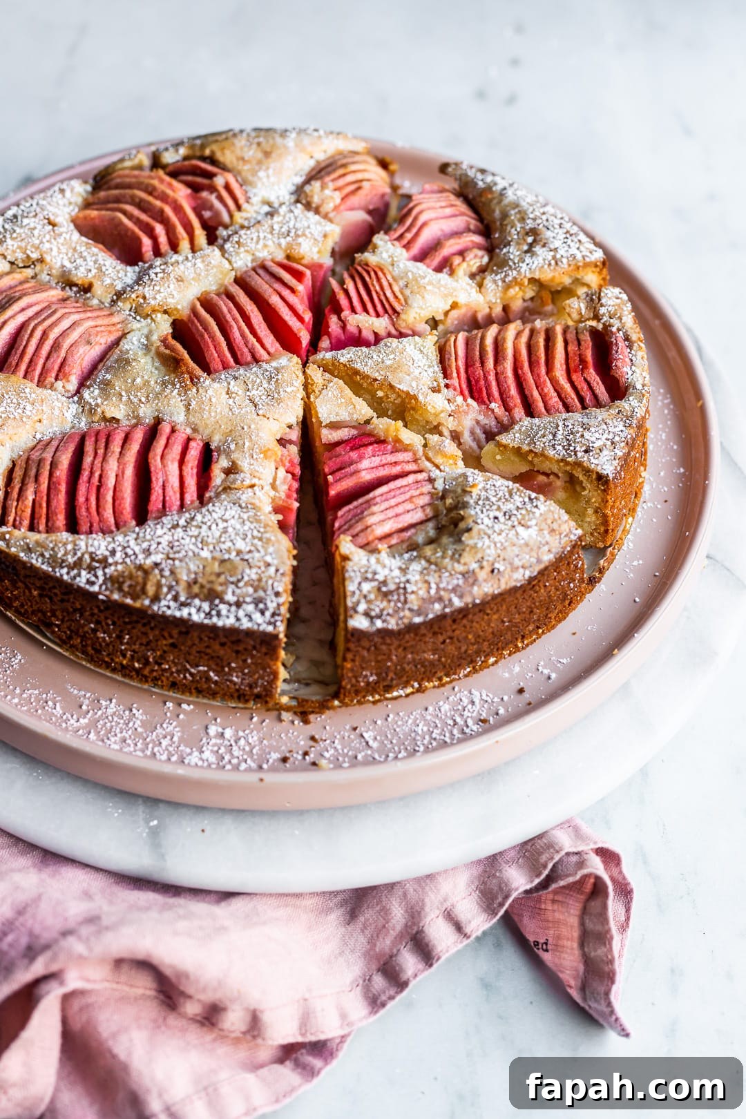 Almond Cake with Roasted Pink Apples 5 Angled view of sliced almond cake with pink apples on a pink plate and marble background