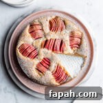 Almond Cake with Roasted Pink Apples 6 A round cake dusted with powdered sugar has a slice cut out of it. It sits on a pink plate atop a white marble platter, which rests on a white marble surface. There are white plates at the top and bottom of the frame.