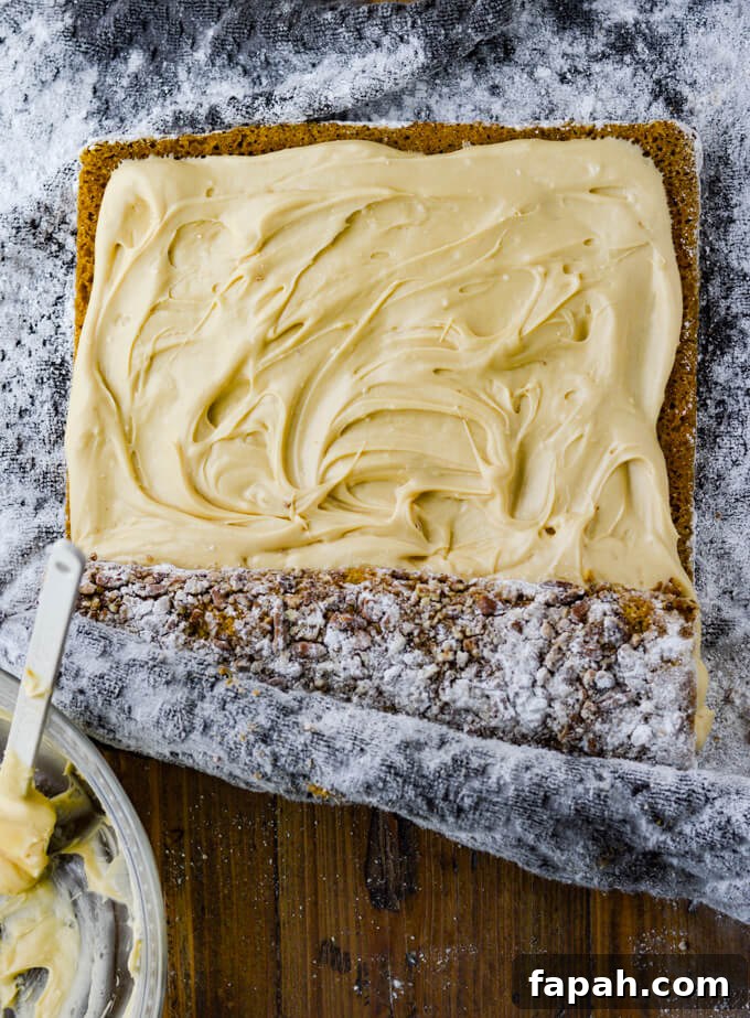 Salted Caramel Pumpkin Cake Roll Recipe for Fall Desserts 11 A frosted pumpkin cake is being rolled up with the help of a dishtowel dusted with powdered sugar. A bowl of empty frosting sits in the corner of the photo.
