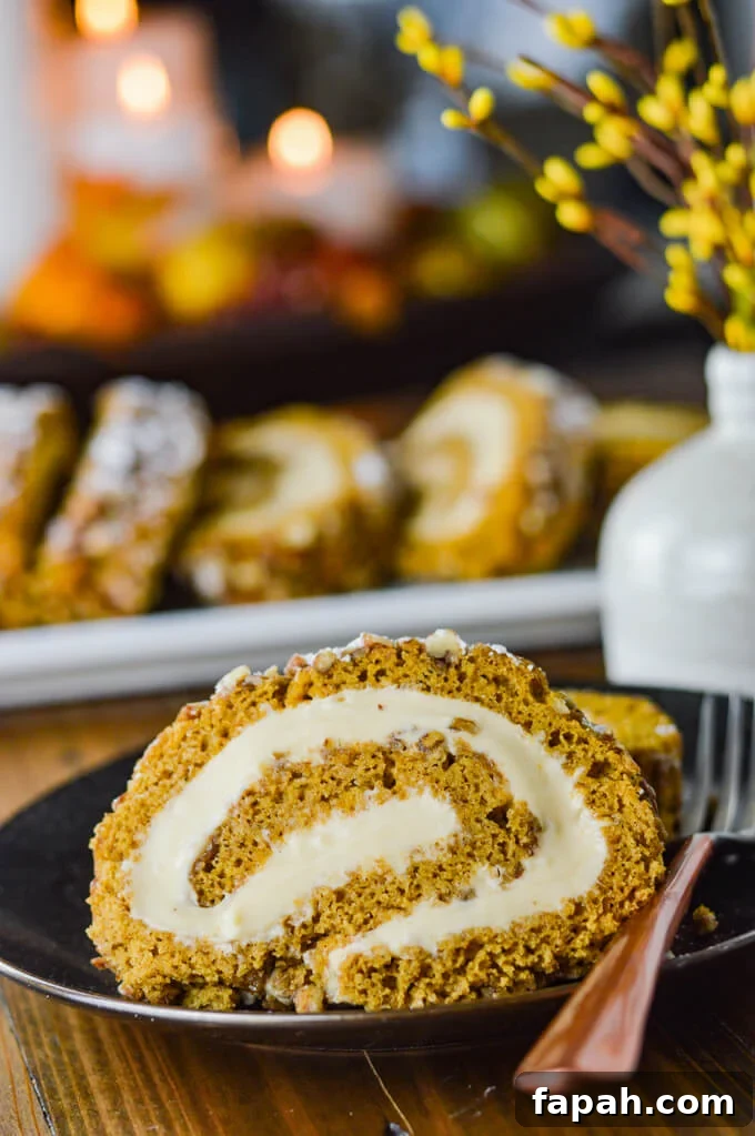 Salted Caramel Pumpkin Cake Roll Recipe for Fall Desserts 12 A close-up of a slice of pumpkin cake roll sits on a black plate with a fork. There's a small white vase with yellow sprigs in it. A pumpkin roll with three slices sit in the background and candles lit in a container in the back is blurred out.