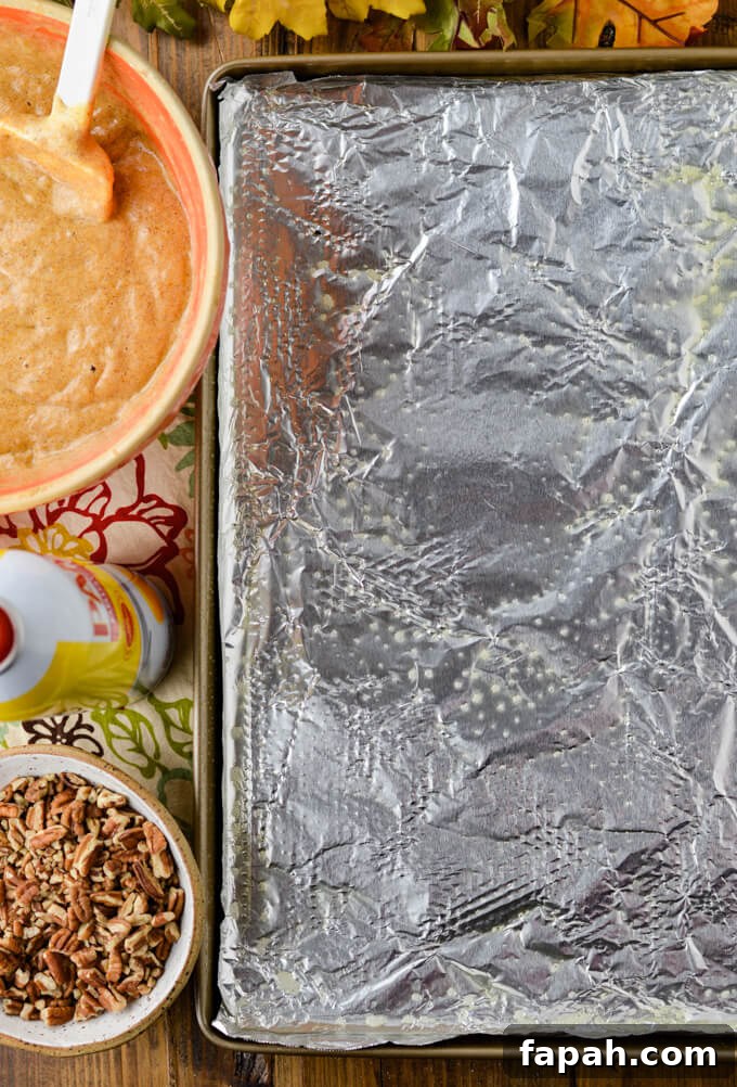 Salted Caramel Pumpkin Cake Roll Recipe for Fall Desserts 6 A foil lined cooking sheet sprayed with Pam with a bowl of pecan pieces, a can of Pam and a bowl of pumpkin cake batter sits next to the pan.