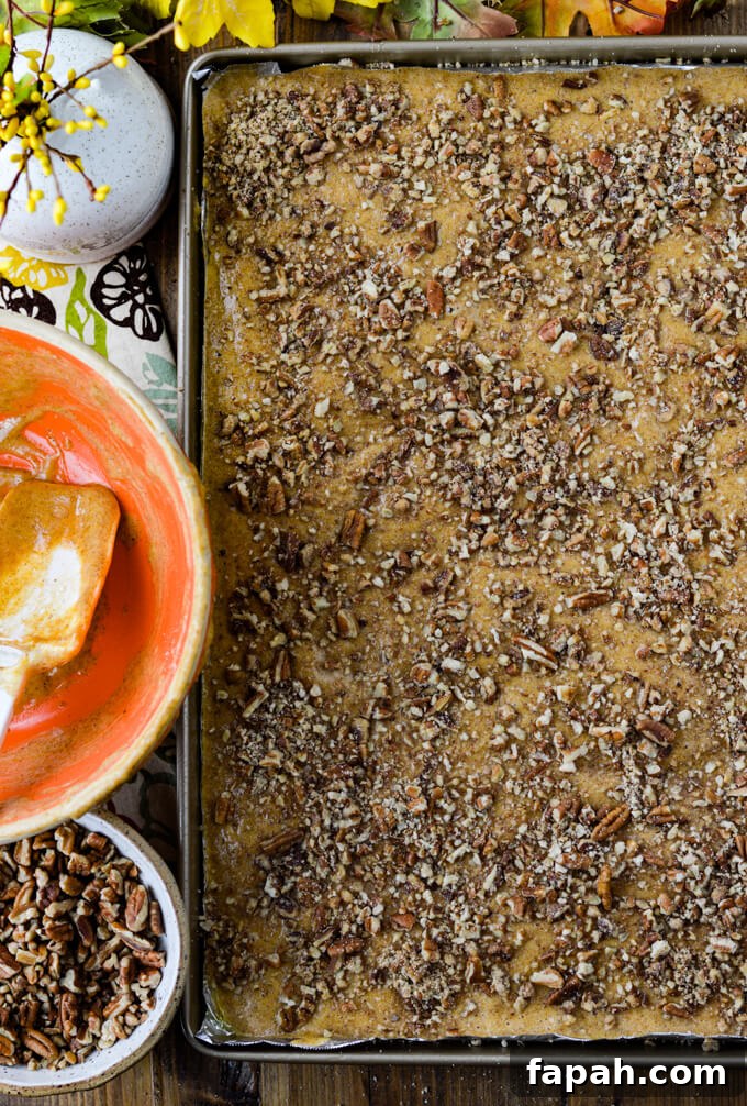 Salted Caramel Pumpkin Cake Roll Recipe for Fall Desserts 7 A cooking sheet with pumpkin cake batter is sprinkled with pecan pieces. A small bowl of pecan pieces and an orange mixing bowl with batter scraped out of it by a white rubber spatula.