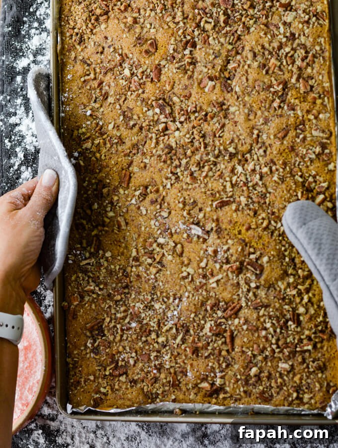 Salted Caramel Pumpkin Cake Roll Recipe for Fall Desserts 8 Baked pumpkin cake sits in a cooking sheet and being held by 2 gray pot holders. It sits above a dishtowel dusted with powder sugar.
