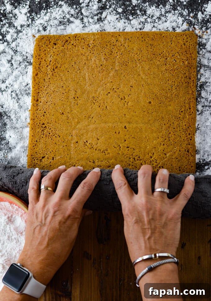 Salted Caramel Pumpkin Cake Roll Recipe for Fall Desserts 9 Two hands are rolling up a baked pumpkin cake in a gray powder sugared dish towel. A small orange bowl holding powder sugar sits in the corner.