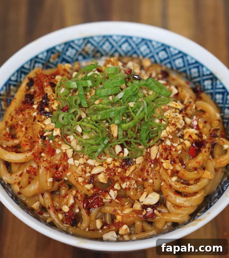 A close-up shot of spicy peanut butter noodles topped with chopped scallions, crushed peanuts, chili flakes, and chili oil, served in a patterned blue and white bowl on a wooden table.