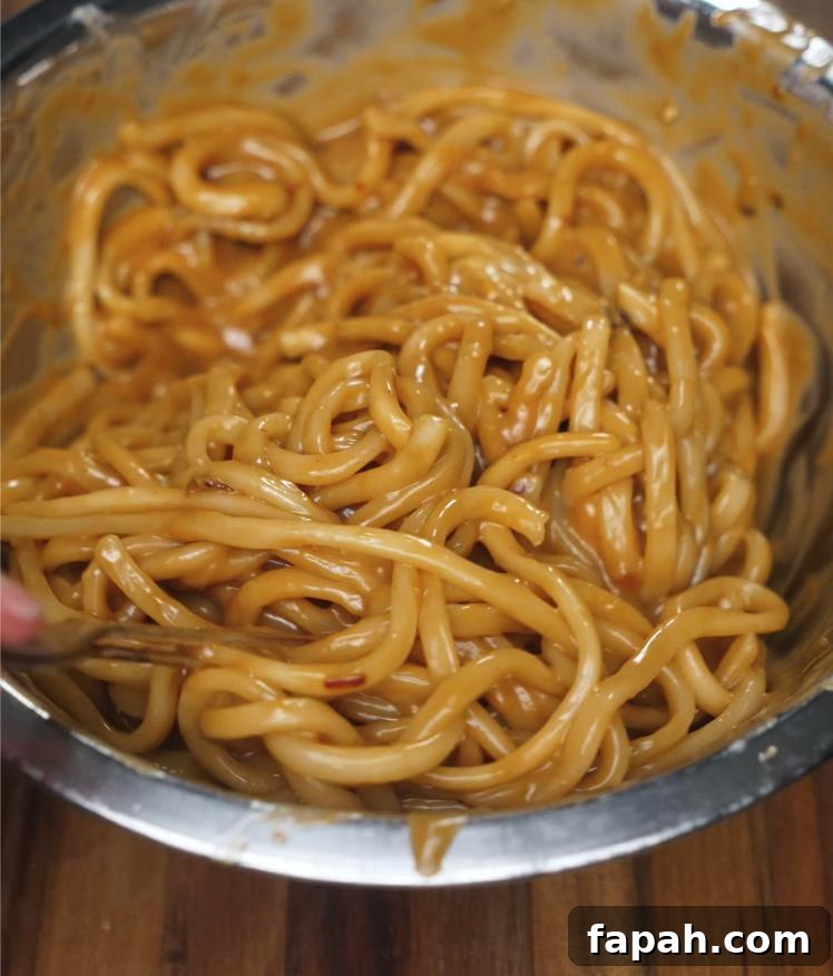 Cooked udon noodles are being mixed with the spicy peanut butter sauce in a large bowl until thoroughly coated, creating a glossy and rich noodle base before garnishing.