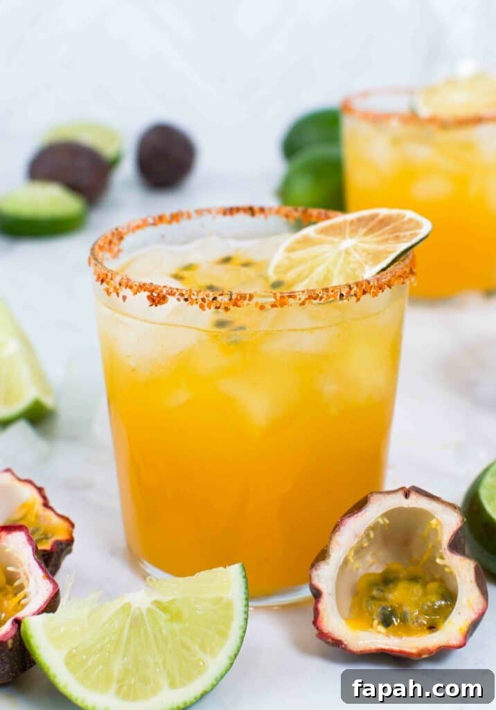 Close up of a passion fruit margarita rimmed with tajin with a dried lime on top on marble surrounded by limes and passion fruits