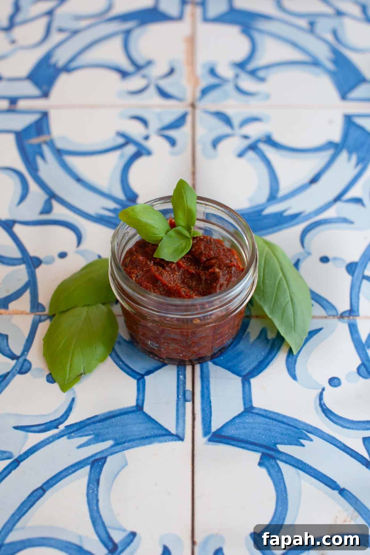 Homemade Pizza Sauce Recipe for Authentic, Restaurant-Quality Flavor 3 A small glass jar of red pesto, perfect as a homemade pizza sauce, garnished with basil leaves and placed on a blue and white patterned tile surface.