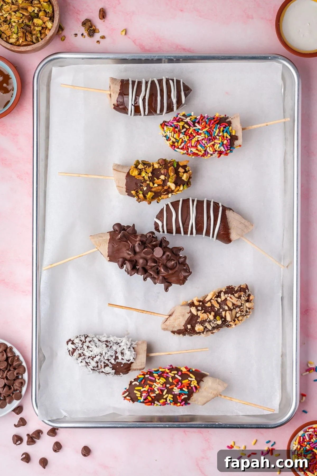 Seven chocolate-covered banana halves on sticks are arranged on a lined baking tray. Each is topped with various toppings, including sprinkles, nuts, coconut, and chocolate drizzle.
