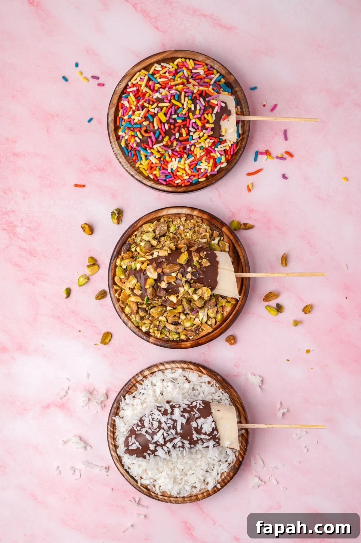 Three chocolate-covered frozen treats are arranged vertically on a pink marble surface. From top to bottom, they're coated with rainbow sprinkles, pistachios, and shredded coconut.