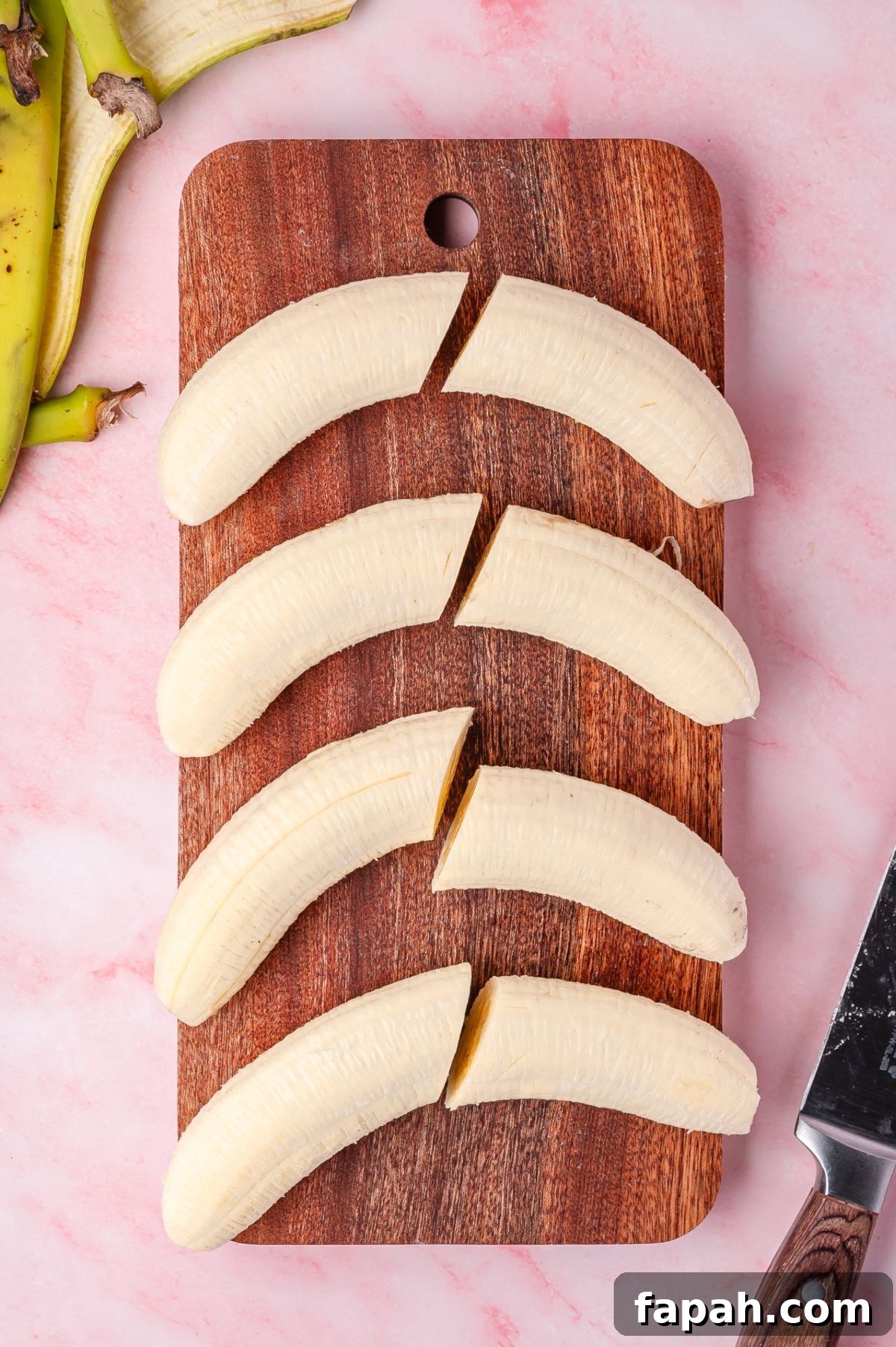 A wooden cutting board with three sliced bananas, each cut into two pieces. A knife and banana peel are nearby on a pink surface.