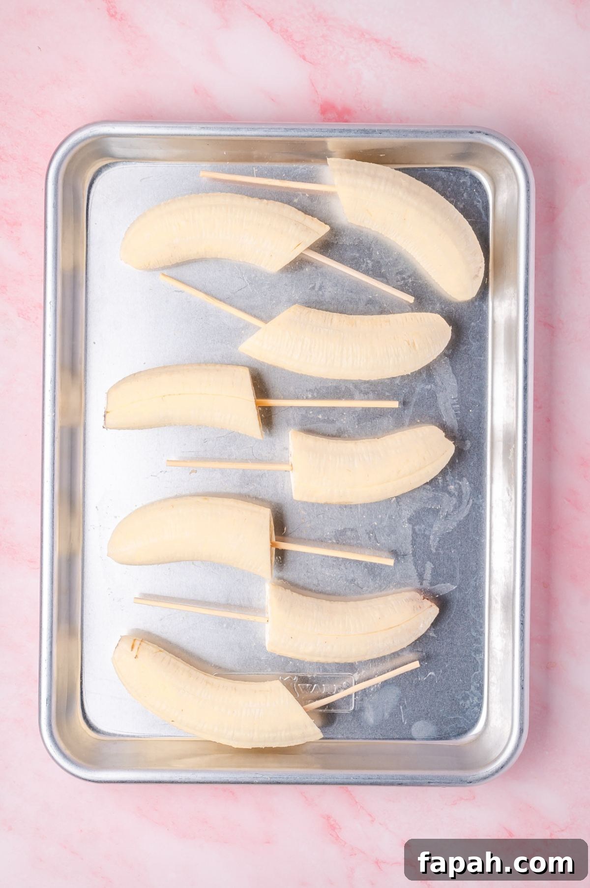 Sliced bananas on skewers are arranged on a metal tray with a pink background.