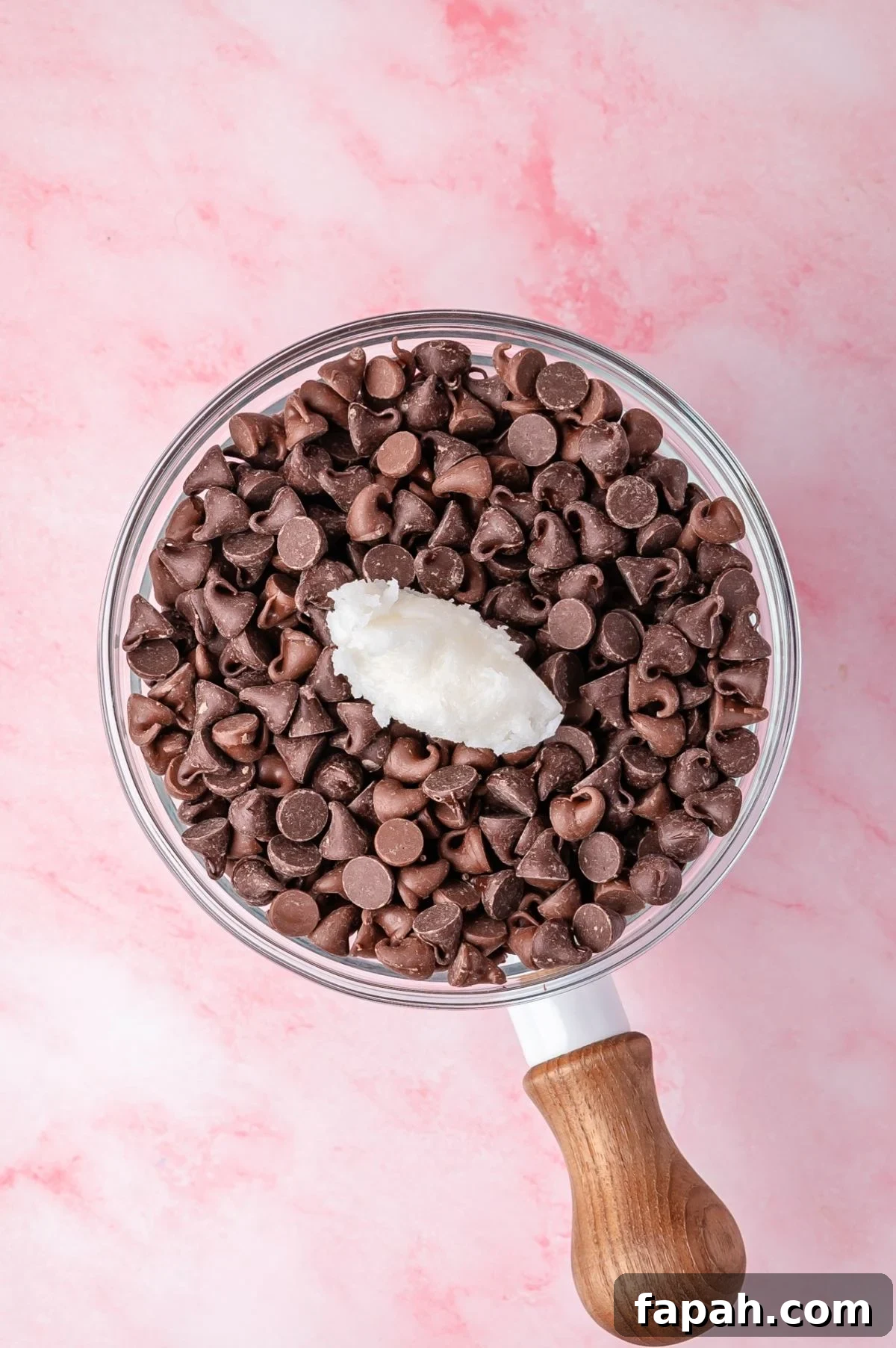 A glass bowl filled with chocolate chips and a dollop of coconut oil, placed on a pink marble surface.