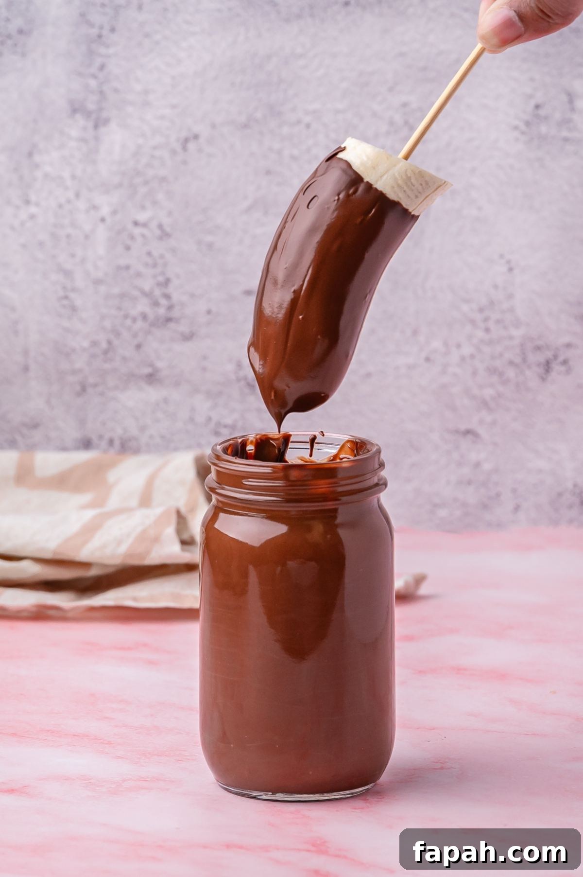 Banana half coated in chocolate held above a jar of melted chocolate.