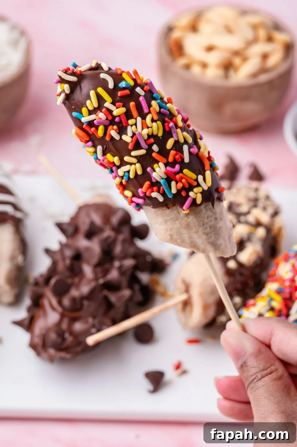 A hand holds a frozen banana covered in chocolate and multicolored sprinkles on a skewer. More chocolate-covered bananas with various toppings are on a plate in the background.