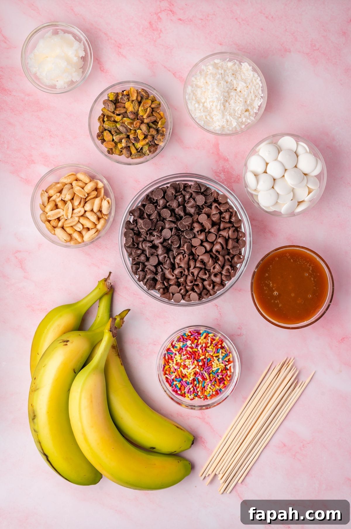 Ingredients for making banana snacks: fresh bananas, chocolate chips, peanuts, pistachios, sprinkles, white candy melts, coconut flakes, caramel sauce, shortening, and skewers on a pink surface.