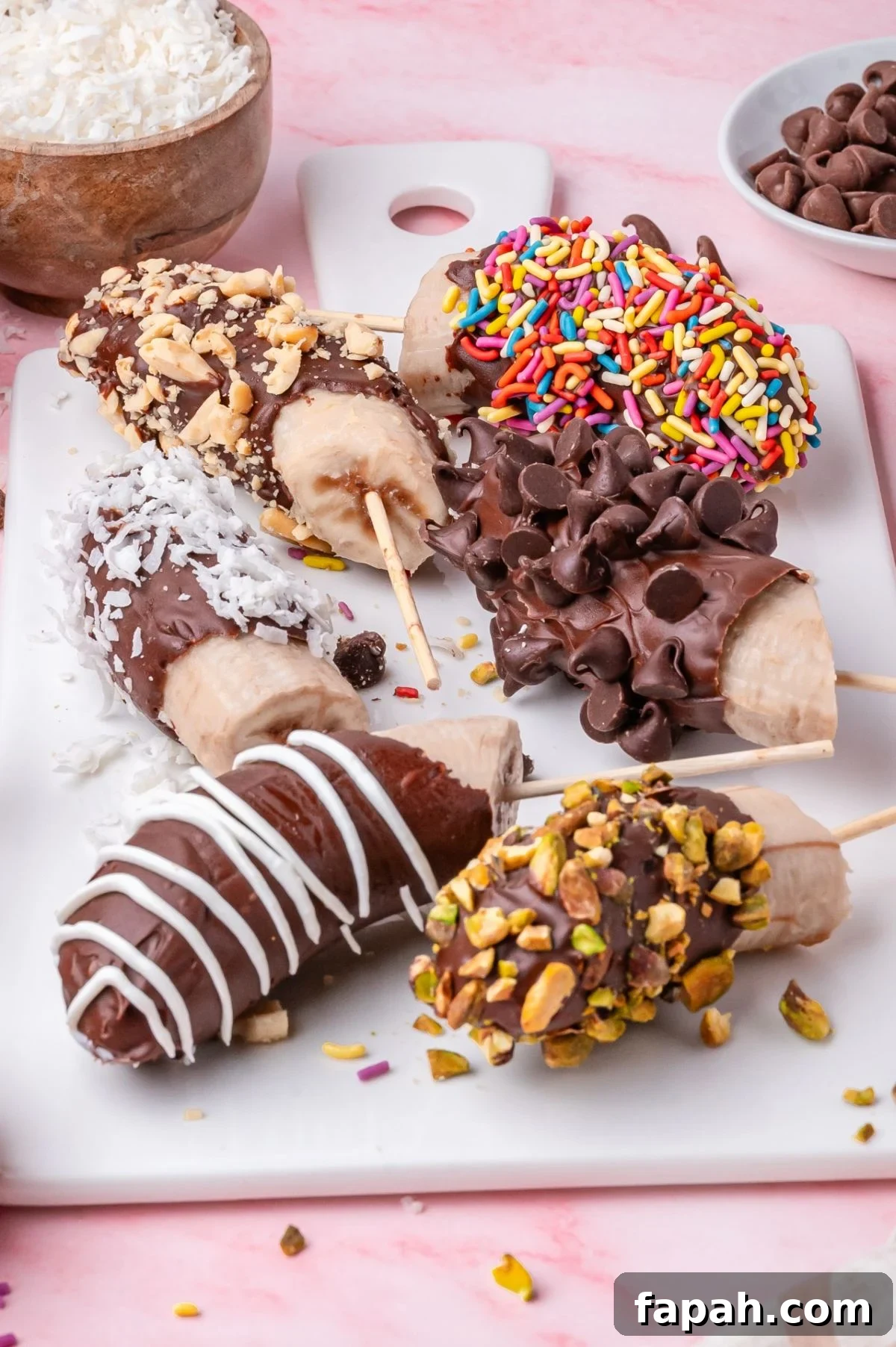 Frozen banana pops coated in chocolate, displayed on a white platter. Toppings include nuts, sprinkles, chocolate chips, coconut, and drizzles. Pink surface beneath the platter.