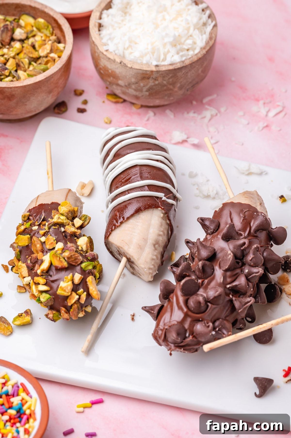 Three chocolate-covered bananas on sticks: one with chopped pistachios, one with white drizzle, and one with chocolate chips, on a white tray, surrounded by bowls of toppings.