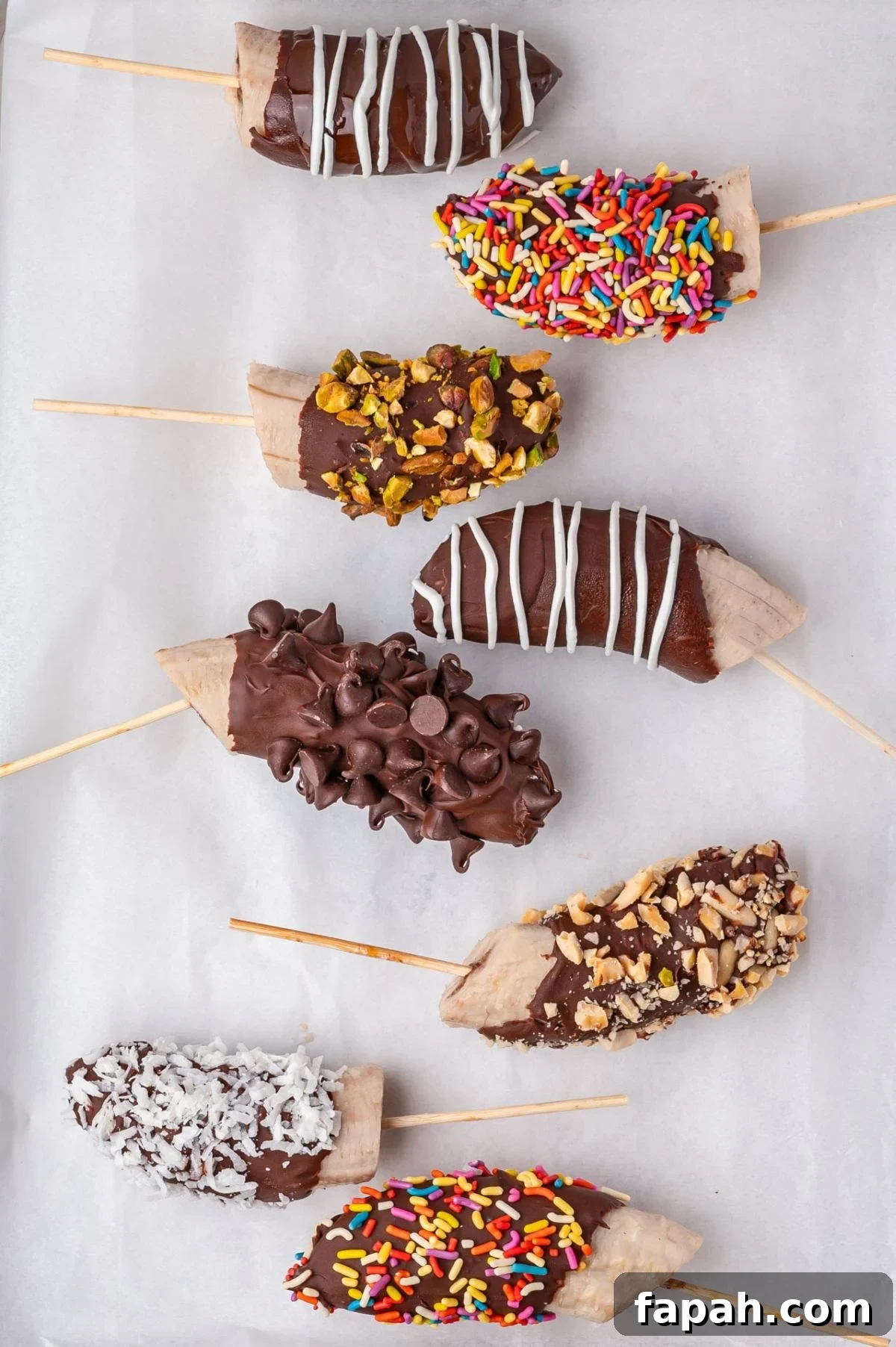 Various chocolate-dipped banana halves on sticks, coated with sprinkles, nuts, or coconut, arranged on parchment paper.