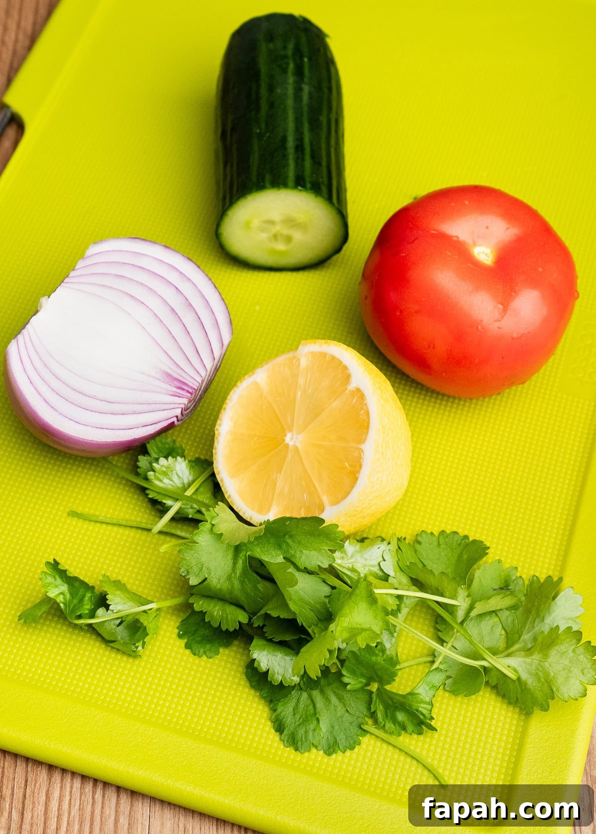 Quarter of cucumber, a tomato, half a red onion, half of a lemon and fresh coriander on a green chopping board
