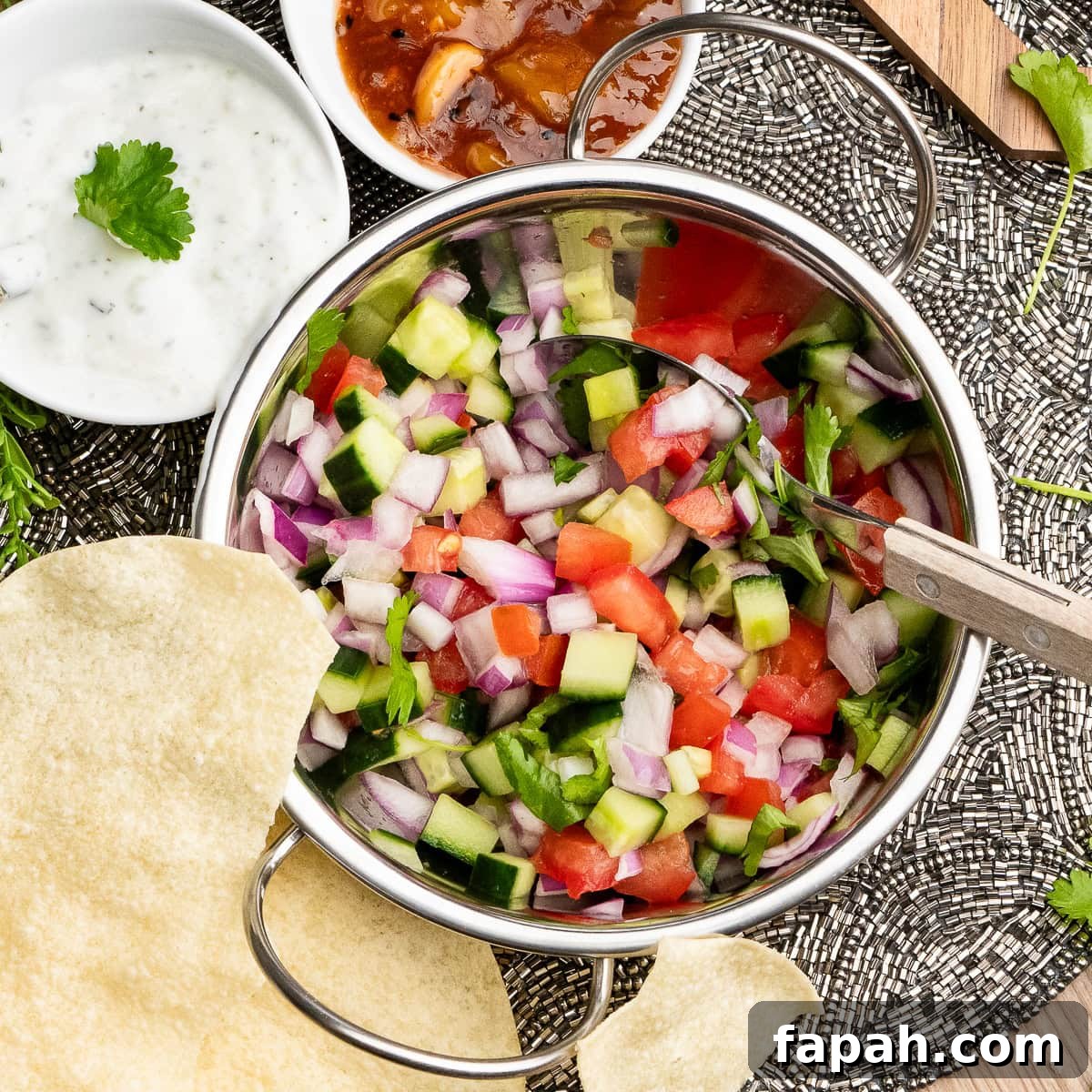 Indian Onion Salad with a spoon on top of the salad in a Balti dish with, mint raita, mango chutney and poppadoms served on the side.