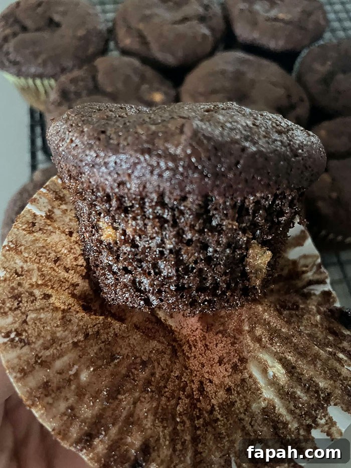A chocolate banana muffin with the muffin paper partially peeled down and more chocolate banana muffins in the background sitting on a cooling rack.