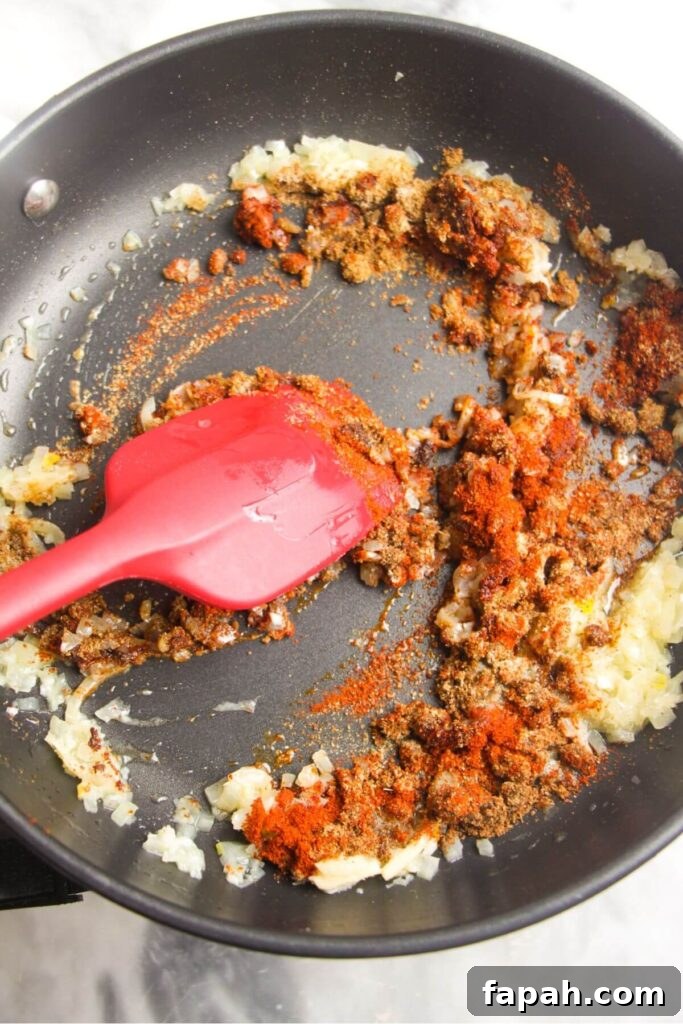 Spices and diced onion stirred by a red spatula in a frying pan.