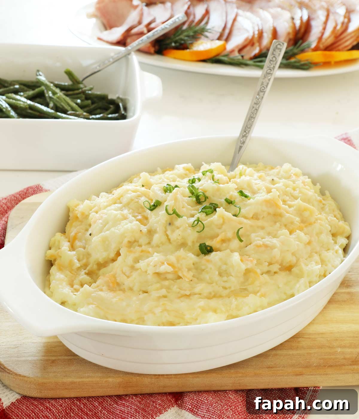 Cheesy mashed potatoes in a white serving dish on a wooden cutting board.