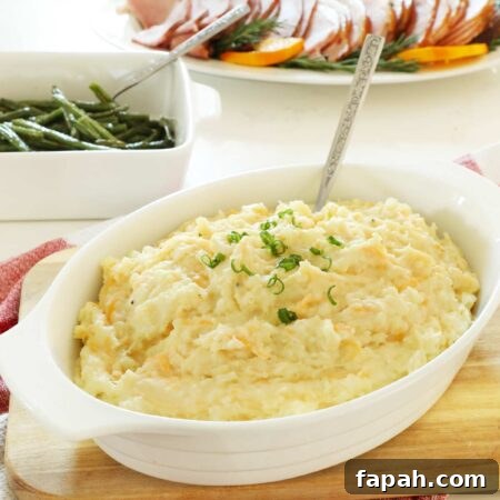 Cheesy mashed potatoes topped with scallions in a white serving dish on a wooden cutting board.