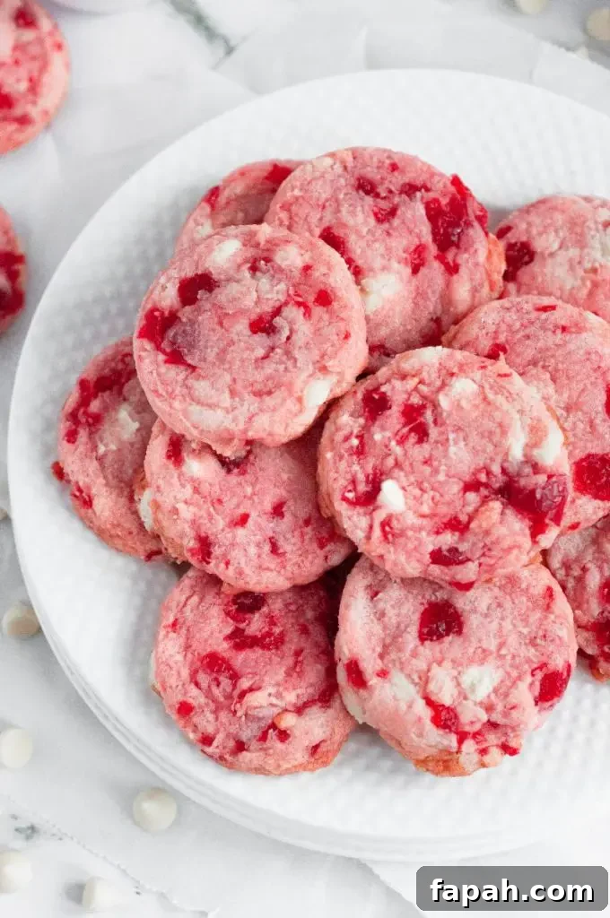 Bright pink cherry chip cookies sitting on a white plate. The cookies have red maraschino cherries in them and white chocolate chips.