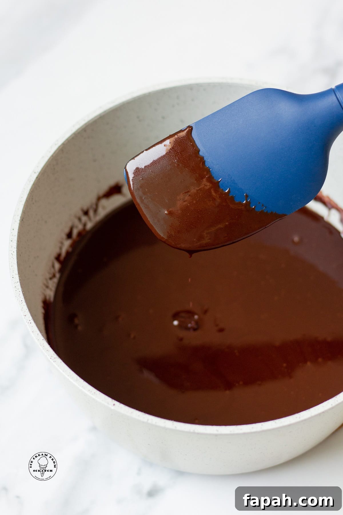 hot fudge sauce in a gray bowl. a blue spatula is holding some up to show the texture and how it sticks to the spoon.