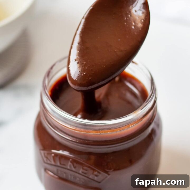 a small glass jar filled with homemade hot fudge. A spoon is lifting some out and the sauce is drizzling back into the jar.