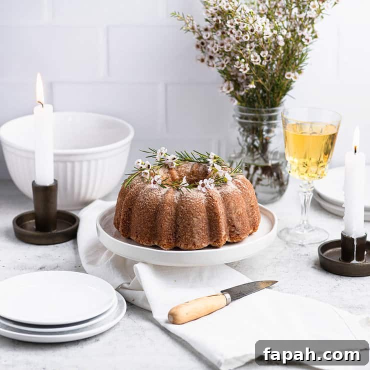 Apple cider donut bundt cake on a white cake stand with a white bowl, plates, candles and a glass of apple cider in the background.