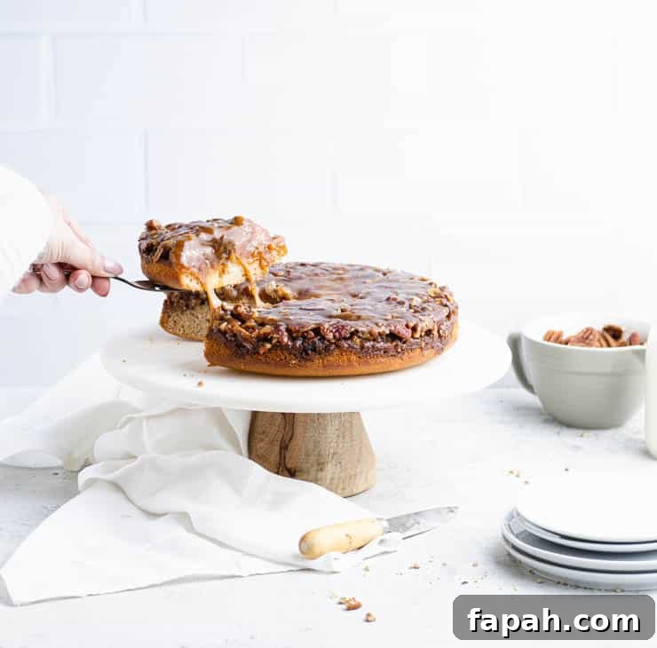 slice being pulled from an pecan upside down cake with bits of caramel pulling between the slice and the rest of the cake with plates and a bowl of pecans in the background.