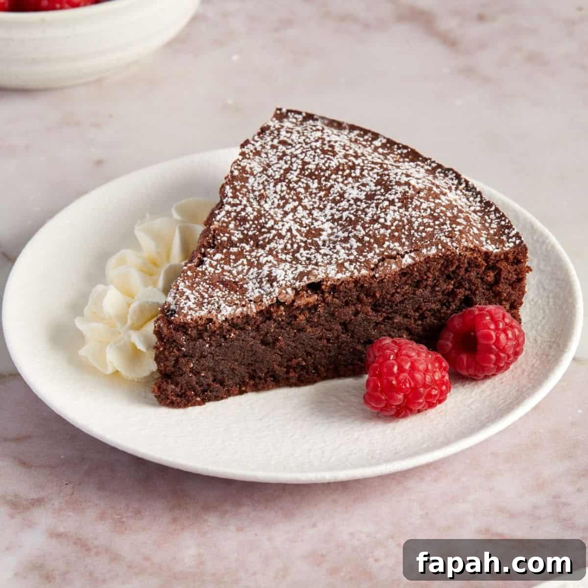 Slice of Torta Caprese, on a small round white plate, with some raspberries and fresh cream on the side.