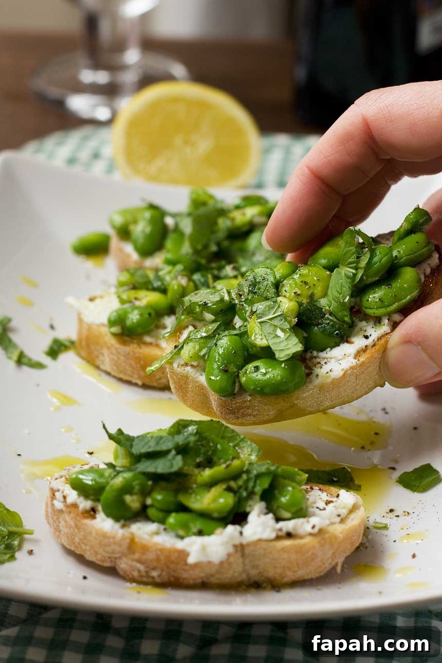 Someone taking a broad bean, mint and ricotta crostini off a plate of 4 with a lemon and checked cloth in the background