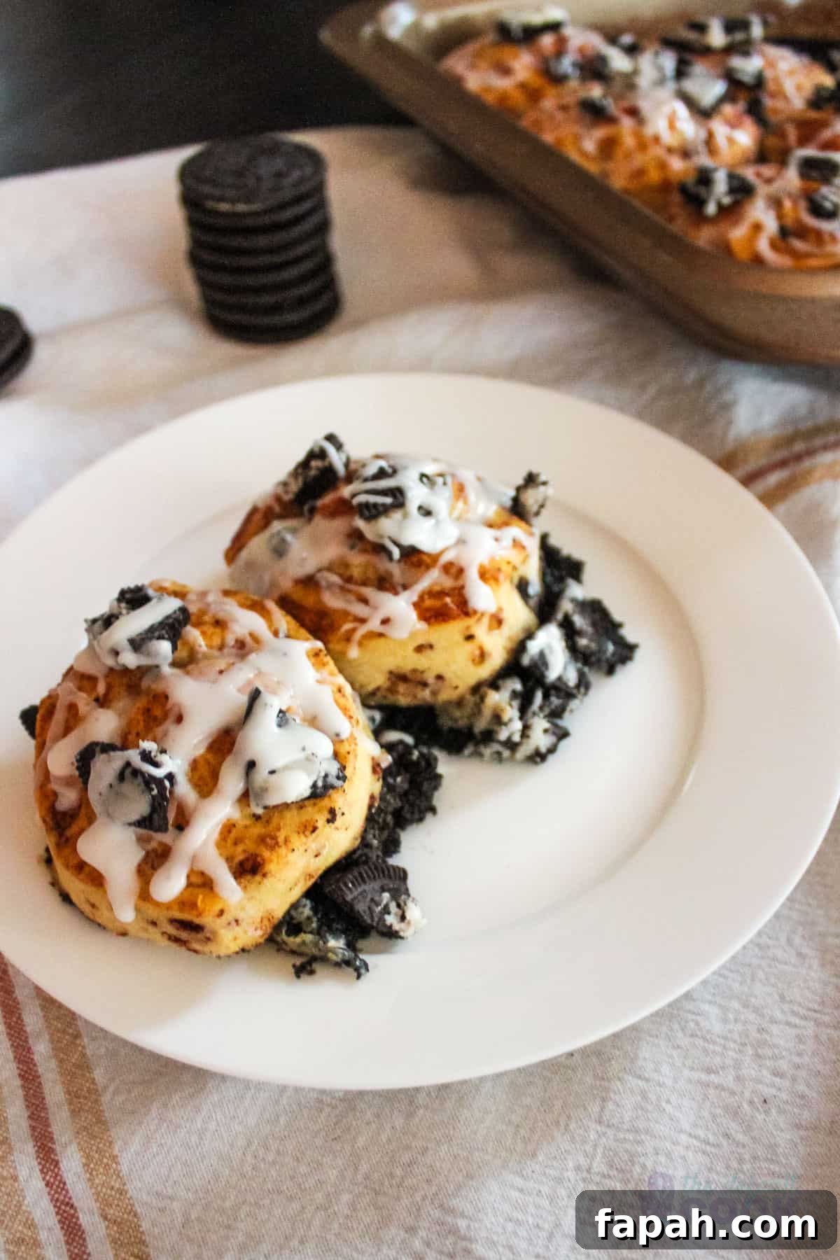 Two oreo cinnamon rolls on a white plate with baking pan and additional oreos in background.