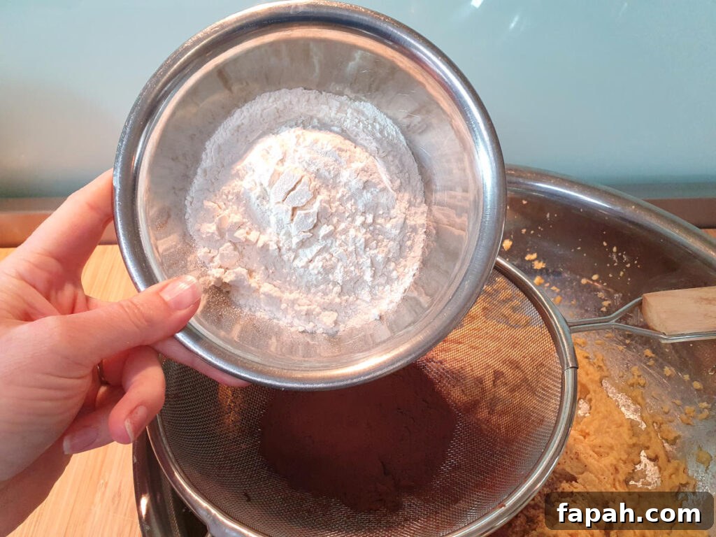 How to Make Homemade Oreo Cookies from Scratch 10 adding flour to other dry ingredients in sieve sittnig on top of mixing bowl with beaten wet ingredients.