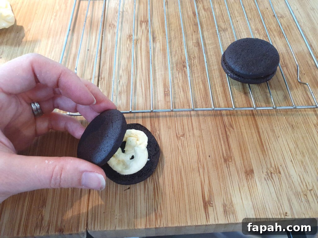 How to Make Homemade Oreo Cookies from Scratch 25 putting top cooking onto bottom cookie with vanilla filling pioped onto it, with write rack and completed fill cookie behind it.