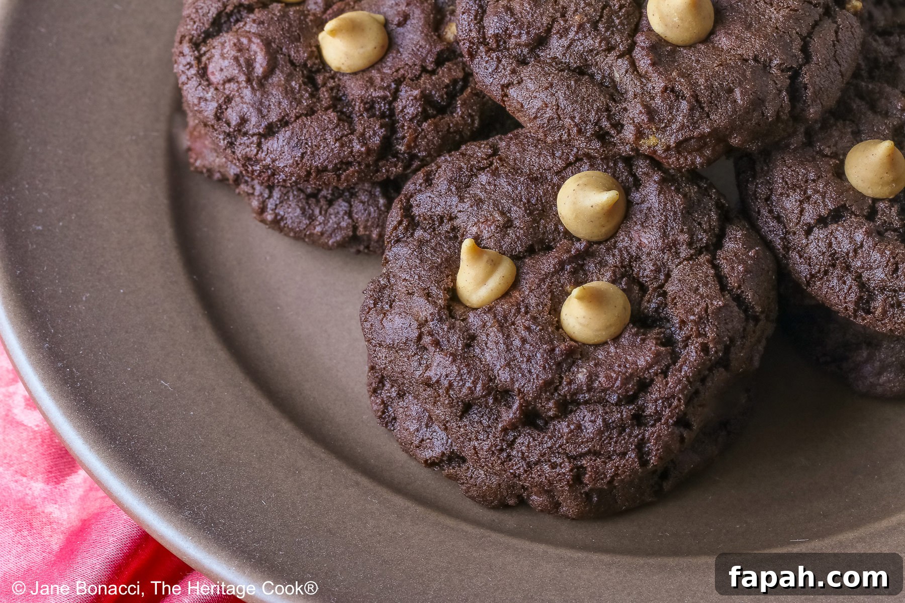 Four stacks of black Double Chocolate Peanut Butter Cookies with light peanut butter chips on top on a deep brown ceramic plate sitting on a bright pink cloth © 2025 Jane Bonacci, The Heritage Cook.